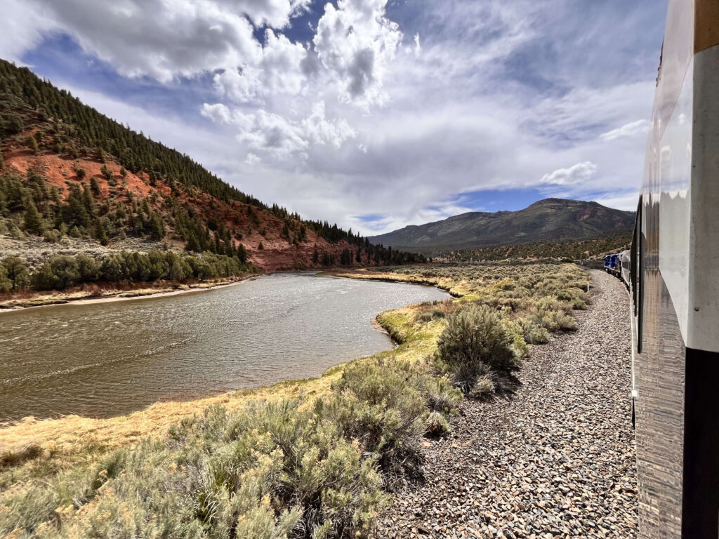Photo taken from the train showing the scene of the Colorado River winding through with a red rock mountain on the side and another mountain in the distance.