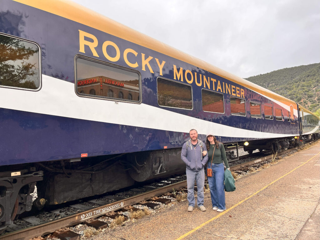 A couple stands together in front of a train with the brand "Rocky Mountaineer" on it. There is a hill with trees in background.