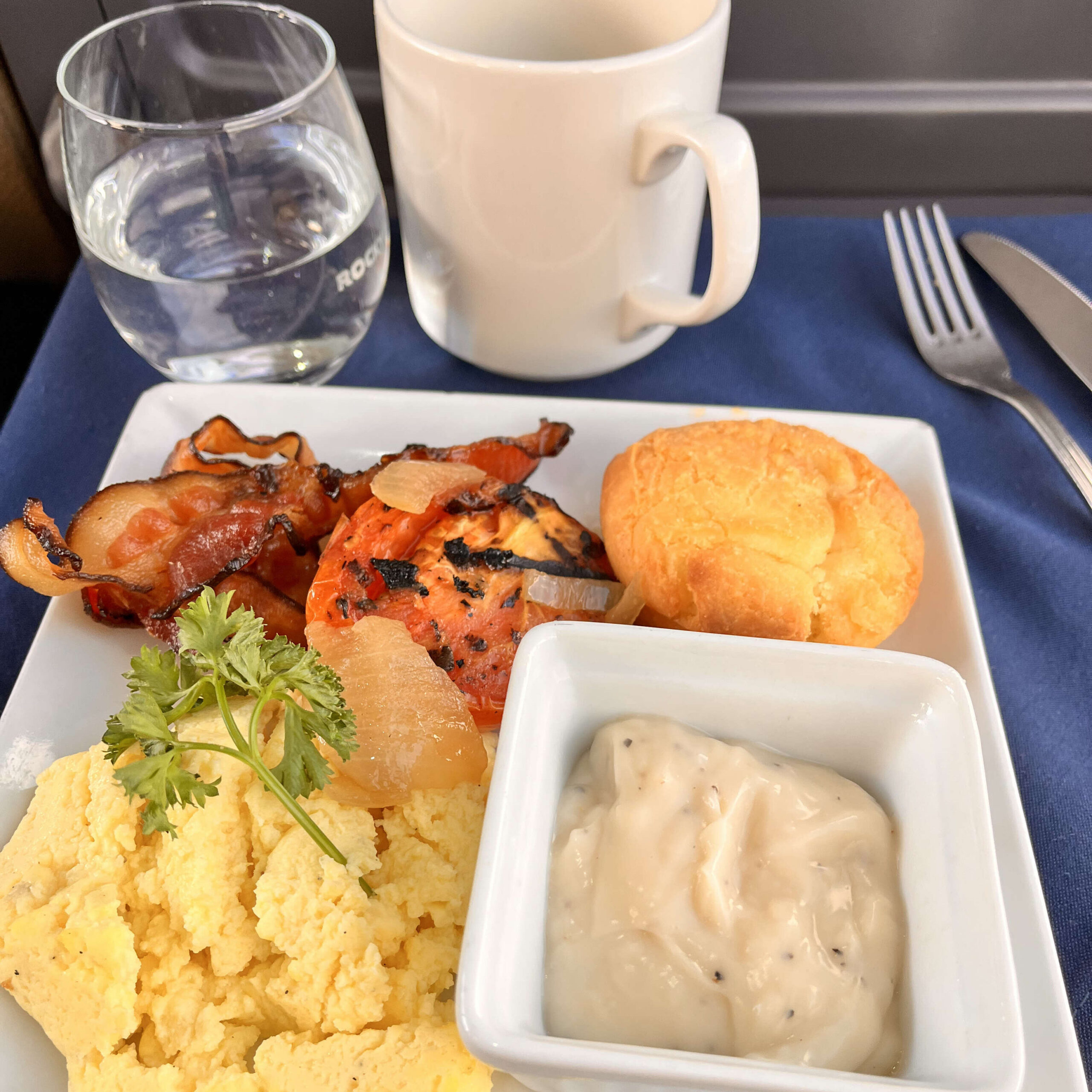 Photo of breakfast on a tray, including a cup of coffee, glass of water, bacon, tomato, scrambled eggs, a gluten free biscuit, and gravy. 