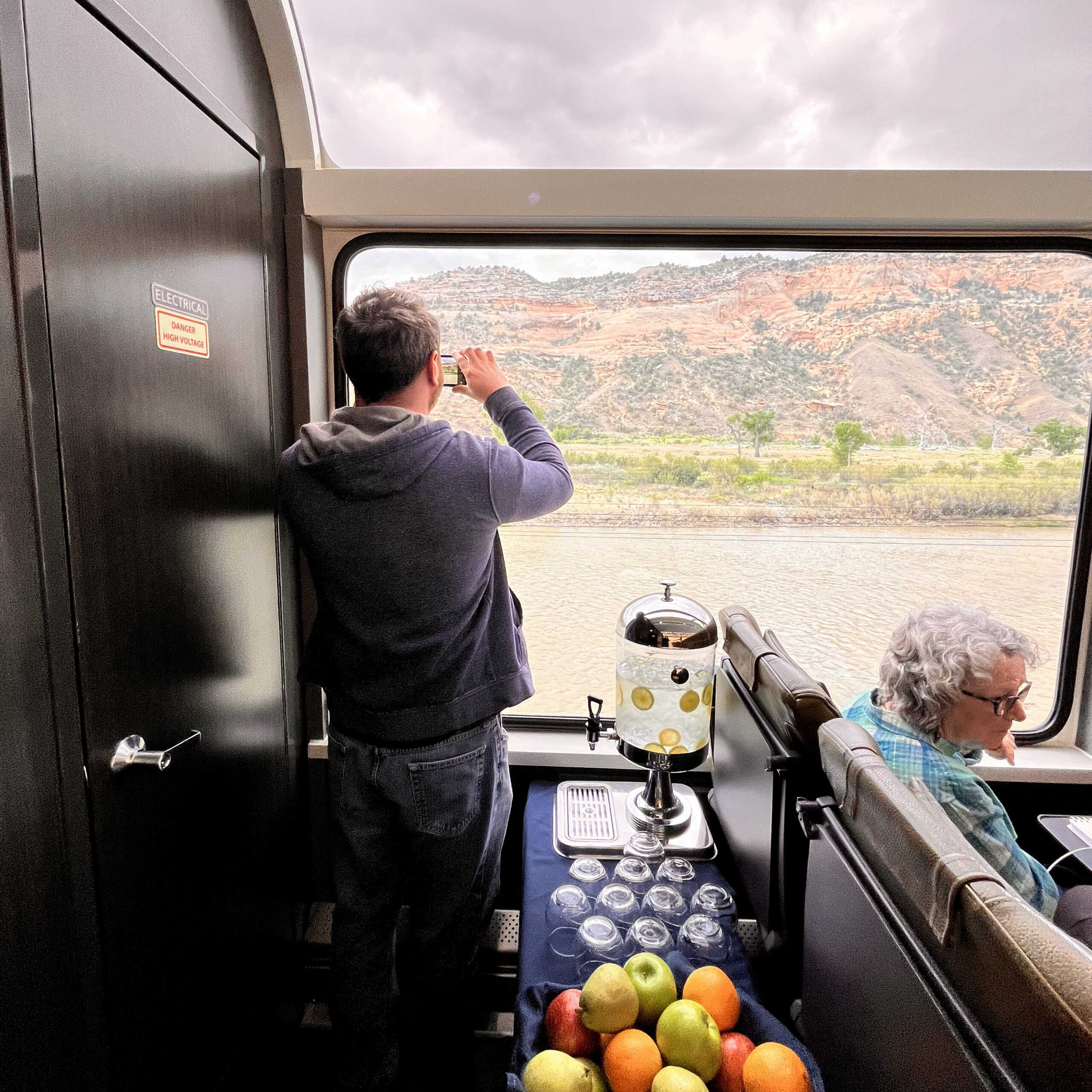 A man stands inside a train looking out the window at the scenery, holding his phone as if he is taking a photo with it. There is a table next to him with fruit, glasses, and a water dispenser.