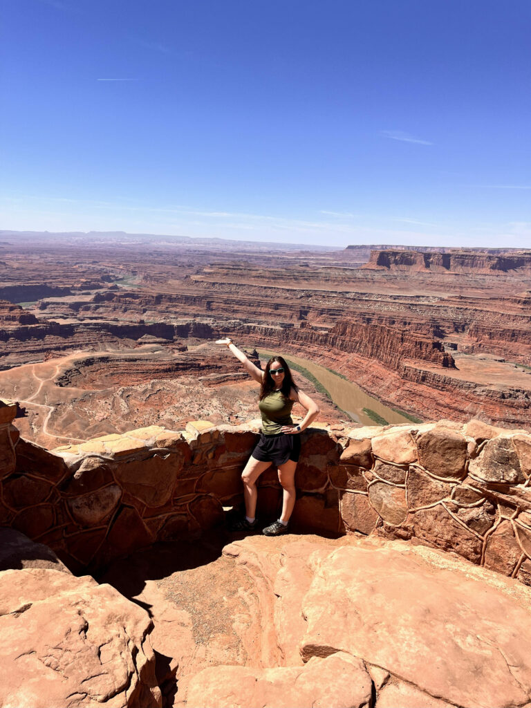 Woman in hiking clothes standing at a stone overlook in Dead Horse Point State Park in Utah, smiling and pointing out over a vast red rock canyon with the Colorado River winding far below under a clear blue sky.
