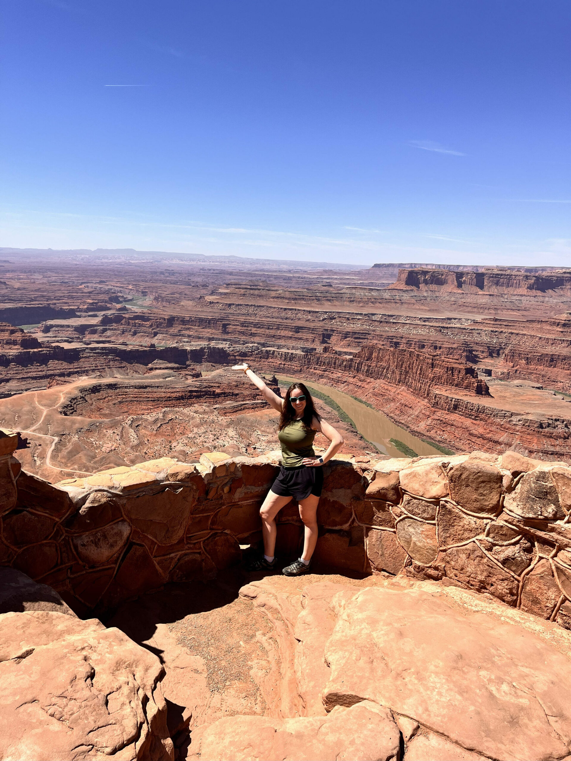Woman in hiking clothes standing at a stone overlook in Dead Horse Point State Park in Utah, smiling and pointing out over a vast red rock canyon with the Colorado River winding far below under a clear blue sky.