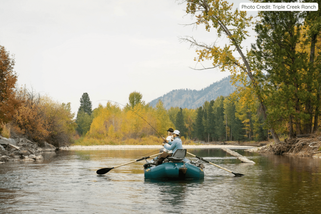 Two people fly fishing from a teal raft on a calm river surrounded by vibrant fall foliage and pine trees at Triple Creek Ranch in Montana’s Bitterroot Mountains.