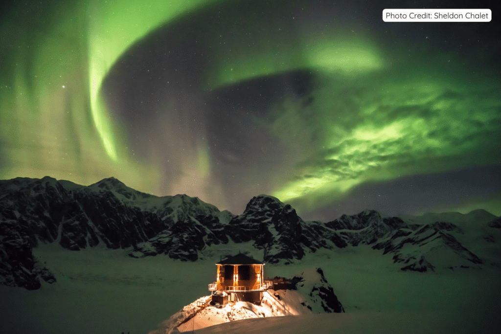 Sheldon Chalet glowing warmly on a snowy ridge in Denali National Park as vibrant green Northern Lights swirl across the starry night sky above the Alaska Range.