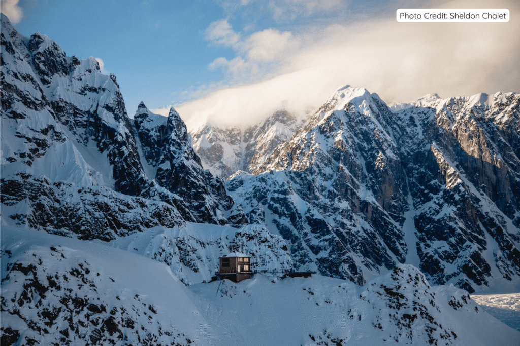 Sheldon Chalet dramatically perched on a rocky outcrop high in the Alaska Range, surrounded by towering snow-covered peaks and dramatic mountain scenery in Denali National Park.