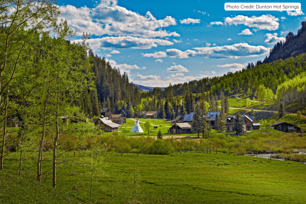 Panoramic view of the restored ghost town at Dunton Hot Springs, with historic cabins, a white tipi, and lush green meadows nestled in a forested valley surrounded by mountains under a bright blue sky.