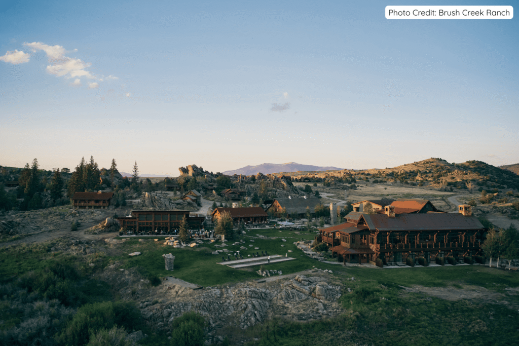 Panoramic view of Brush Creek Ranch’s expansive lodge buildings and grounds nestled among rocky outcrops and open Wyoming landscape at golden hour, with guests gathered outdoors under a clear blue sky.