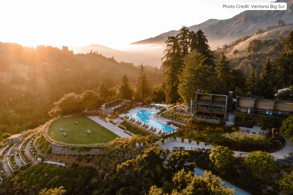 Golden sunrise light bathing Ventana Big Sur’s infinity pool, lawns, and modern wooden buildings nestled among redwood trees and rolling hills, with mist lingering in the valleys below.