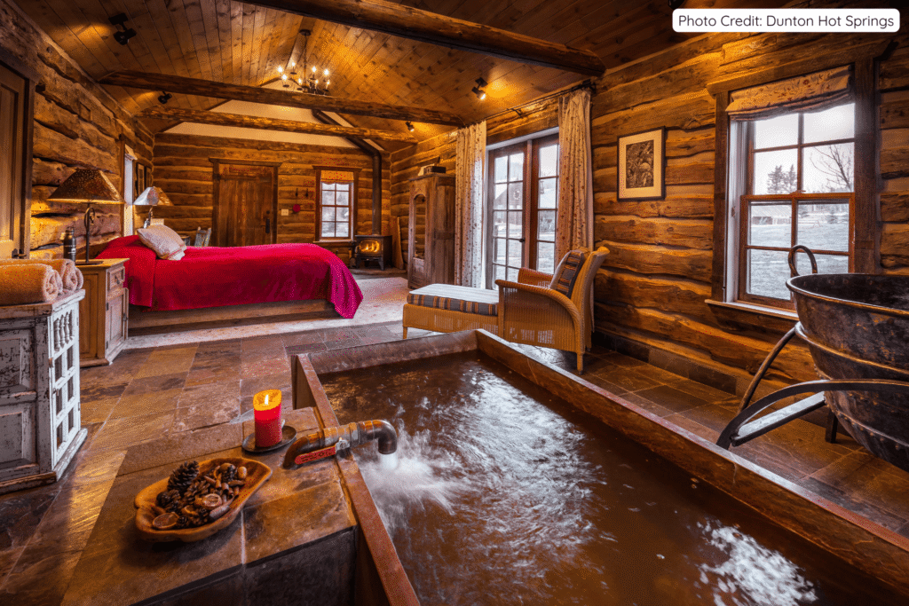 Cozy log cabin interior at Dunton Hot Springs featuring a large wooden soaking tub with flowing mineral water, a vibrant red bed, warm lighting, and rustic wooden beams throughout the room.
