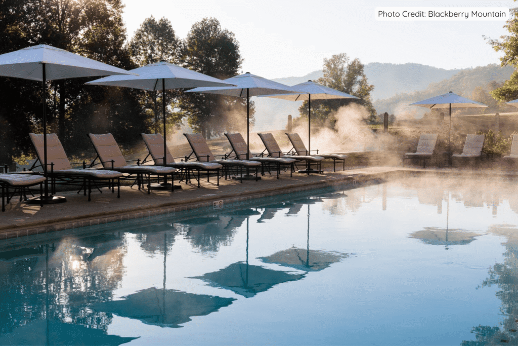 A serene infinity pool at Blackberry Mountain with steam gently rising in the soft morning light, surrounded by lounge chairs under white umbrellas and misty forested hills in the background.