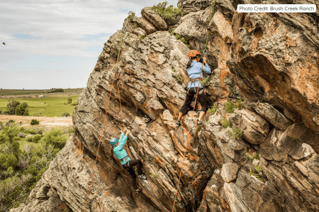 Two climbers with helmets and harnesses scaling a rugged rock face at Brush Creek Ranch, with one in a blue jacket higher up and another in a turquoise top below, overlooking green fields and open countryside.