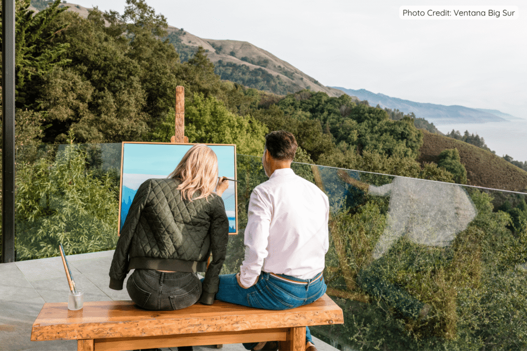 A couple sitting on a wooden bench painting on an easel overlooking the dramatic hills and coastline of Big Sur at Ventana Big Sur, with one person in a green jacket actively painting while the other watches.