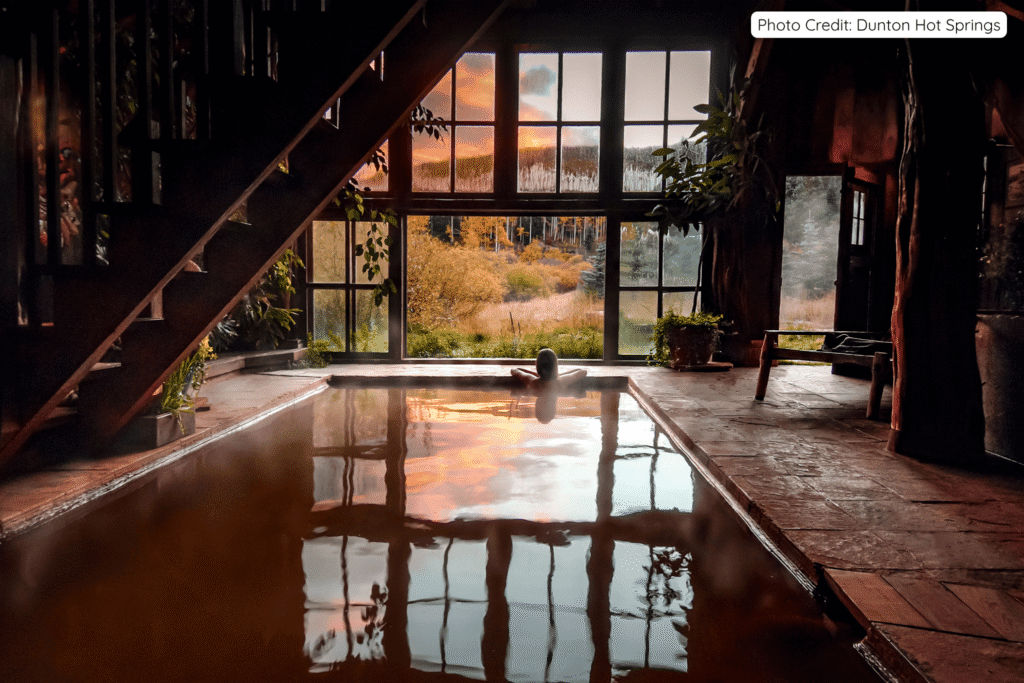 A person relaxing in a steaming indoor hot springs pool inside a rustic wooden bathhouse at Dunton Hot Springs, with a dramatic sunset glowing through large windows overlooking the Colorado mountains.