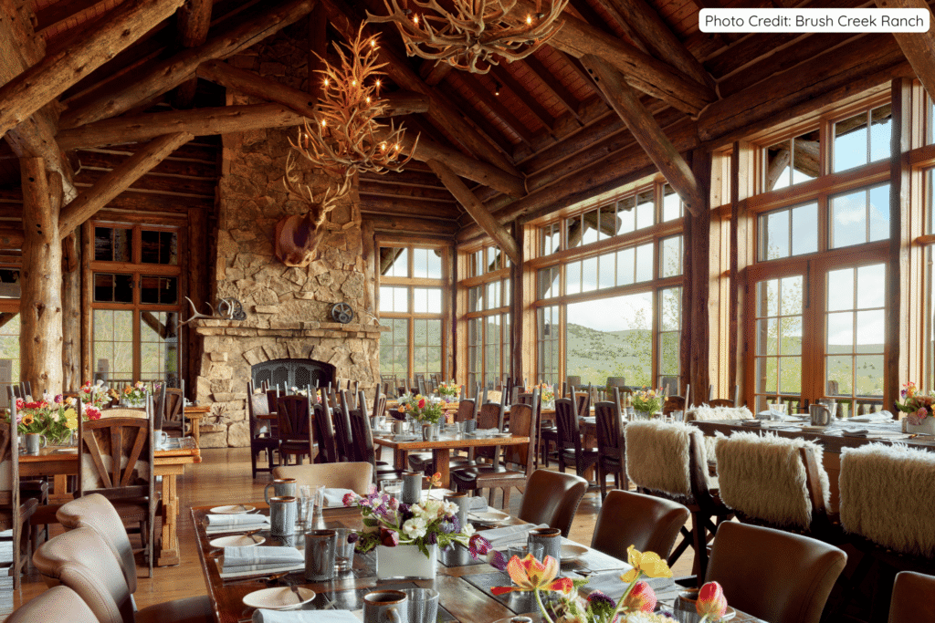 Elegant rustic dining room at Brush Creek Ranch featuring massive wooden beams, a stone fireplace with a mounted elk head, long wooden tables set with flowers, and glowing antler chandeliers hanging from the ceiling.