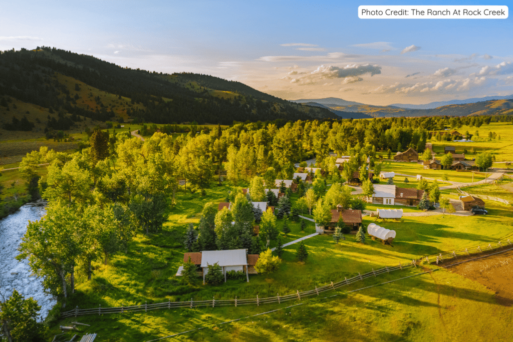 Aerial view of The Ranch at Rock Creek in Montana, showing luxury cabins, covered wagons, lush green meadows, a winding river, and forested hills under a bright blue sky at golden hour.