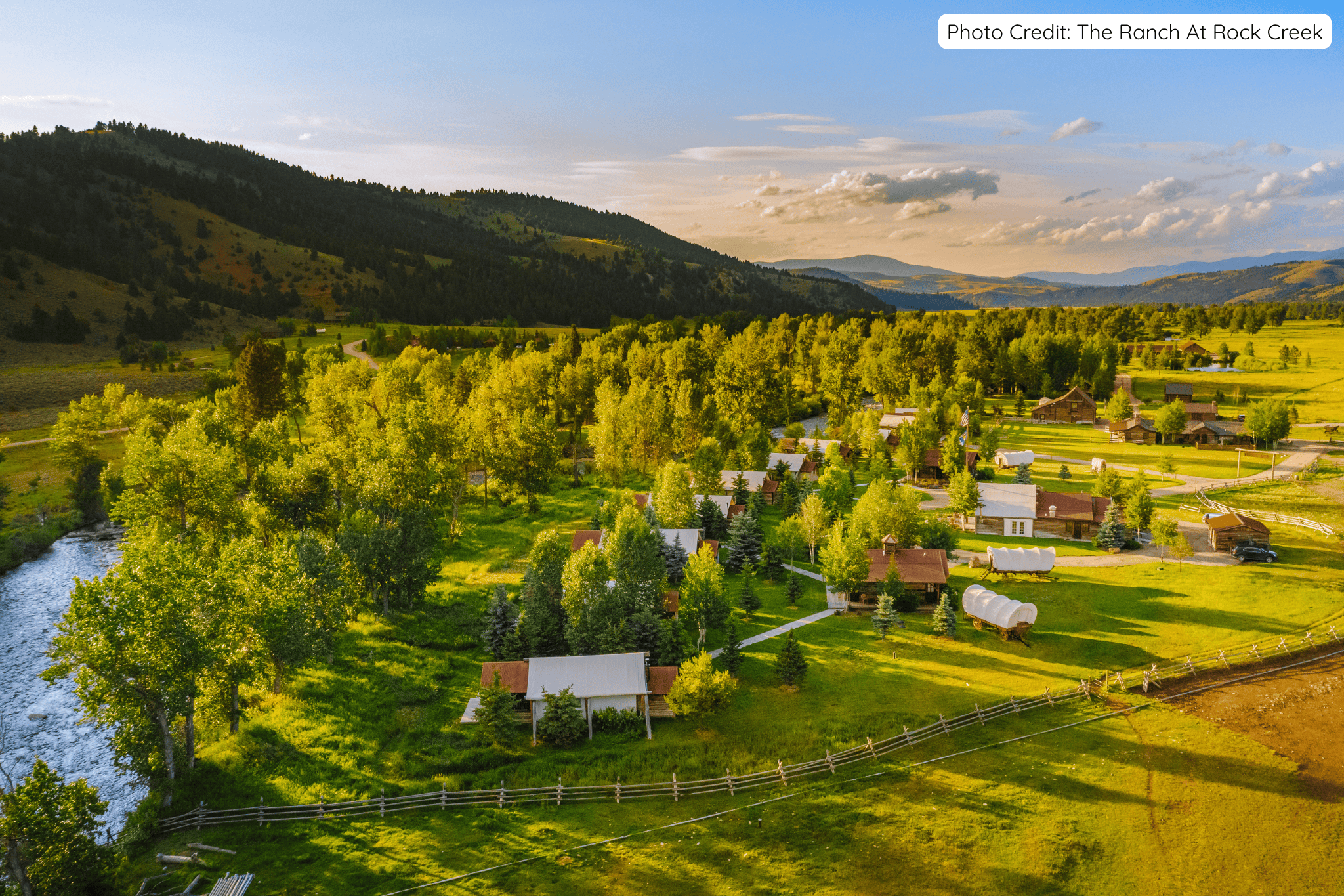 Aerial view of The Ranch at Rock Creek in Montana, showing luxury cabins, covered wagons, lush green meadows, a winding river, and forested hills under a bright blue sky at golden hour.