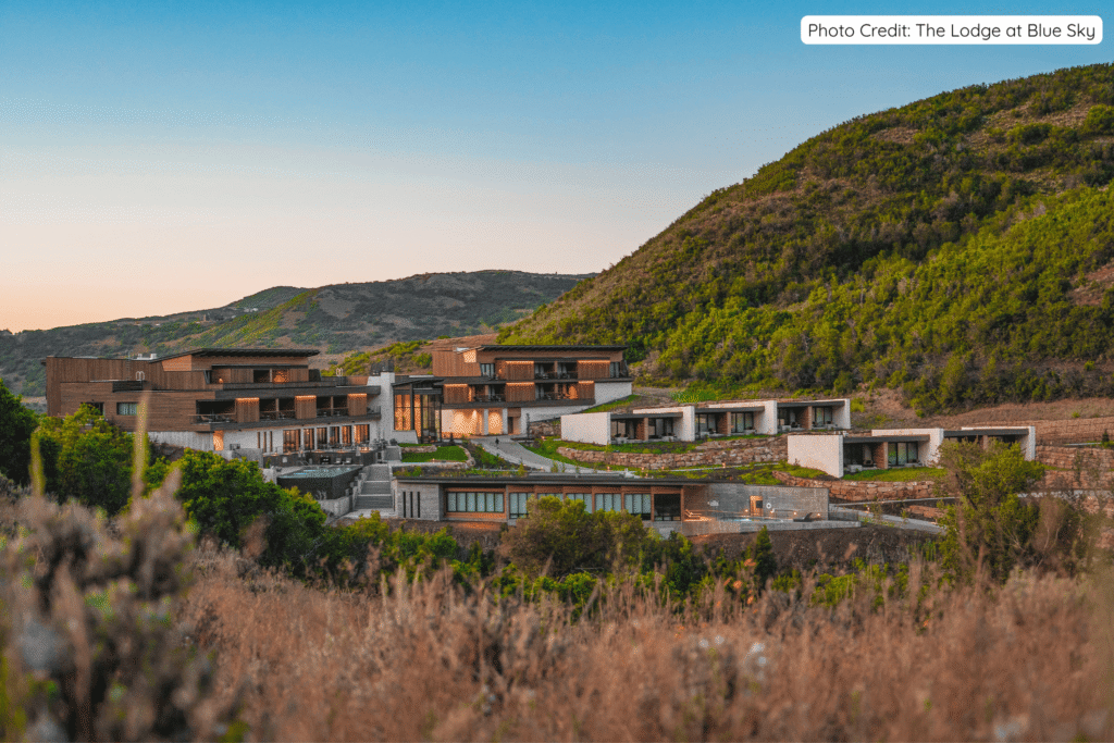 Modern luxury lodge buildings with warm lighting nestled into rolling hills and green landscape at Brush Creek Ranch in Wyoming, with expansive ranch terrain stretching into the distance at sunset.