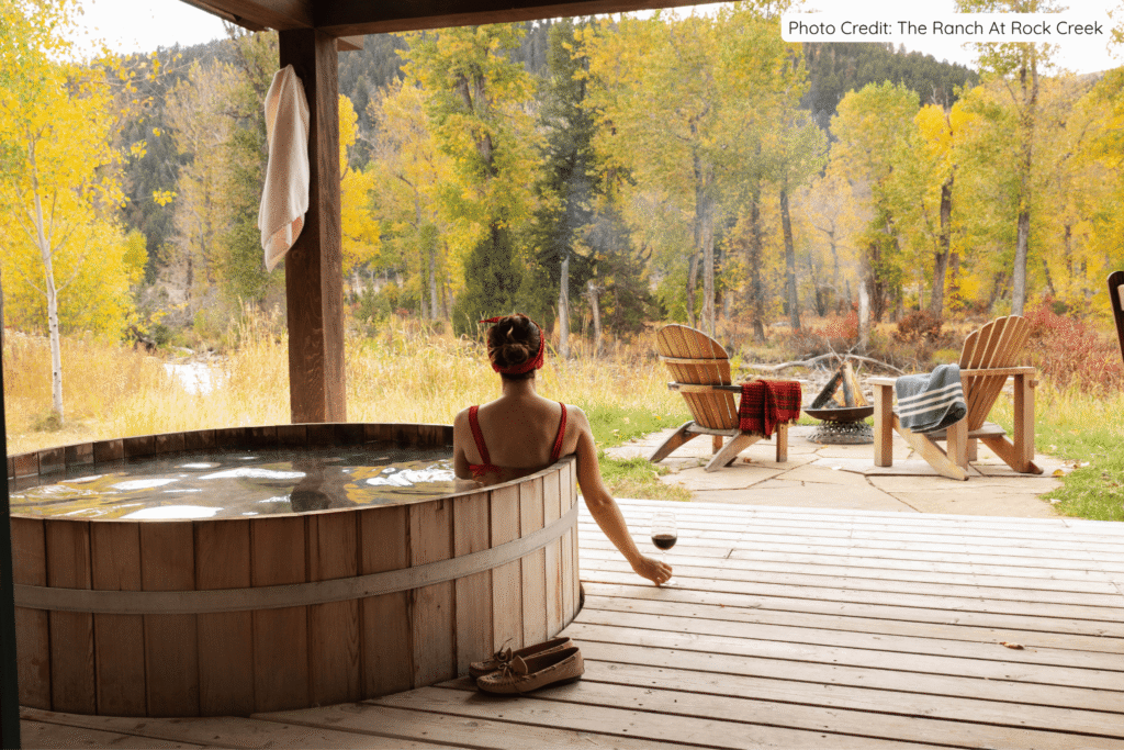 A woman relaxing in a steaming wooden hot tub on a deck at The Ranch at Rock Creek, enjoying a glass of wine while overlooking colorful autumn aspen trees and a forested mountain backdrop in Montana.