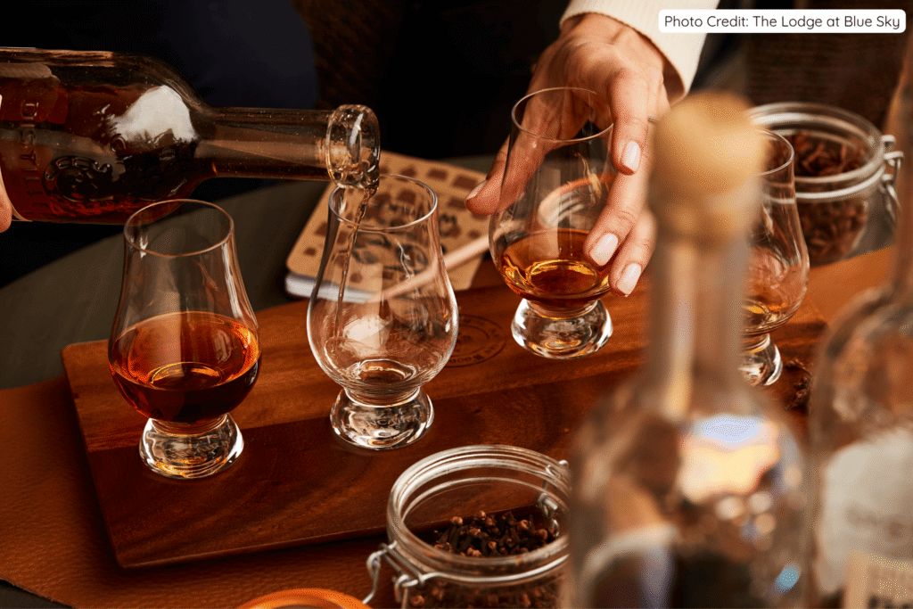 A close-up of amber whiskey being poured from a glass bottle into tasting glasses on a wooden board, with hands holding a glass and jars of spices nearby, capturing an intimate tasting experience.