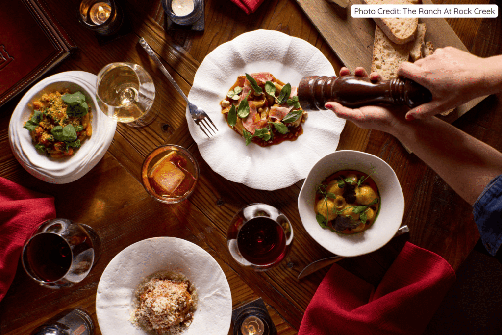 Top-down view of an elegant wooden dining table at The Ranch at Rock Creek, featuring gourmet dishes like pasta with prosciutto, fresh salads, wine glasses, and hands grinding fresh pepper over a plated meal.