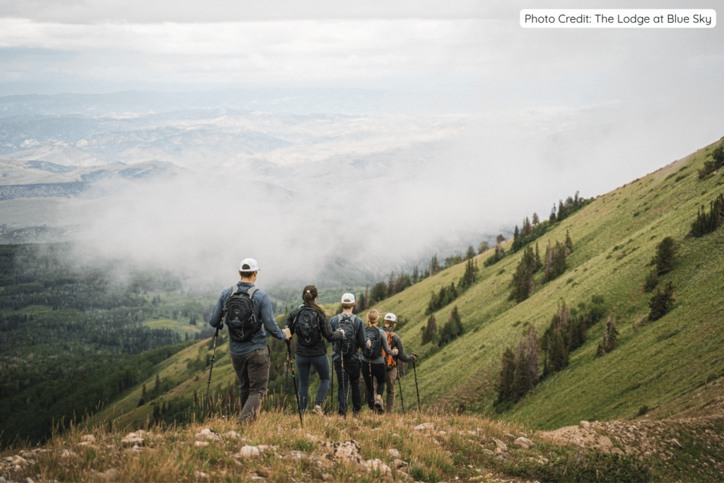 A group of hikers with backpacks and trekking poles standing on a grassy ridge, looking out over vast misty valleys and mountain ranges during a guided hike at Brush Creek Ranch.