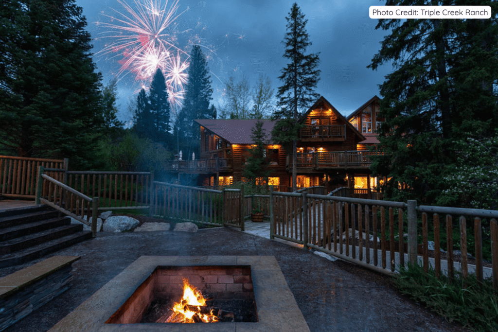 A glowing fire pit in the foreground with bright fireworks exploding in the night sky above the illuminated log lodges and tall pine trees at Triple Creek Ranch.